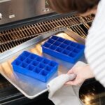 A person placing two silicone ice cube trays on a baking pan into an oven.